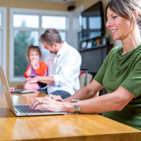 A woman working on a laptop at her kitchen table with her husband and daughter in the background.