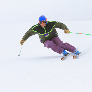 Male downhill skier wearing purple pants and a blue helmet skiing down a groomed slope