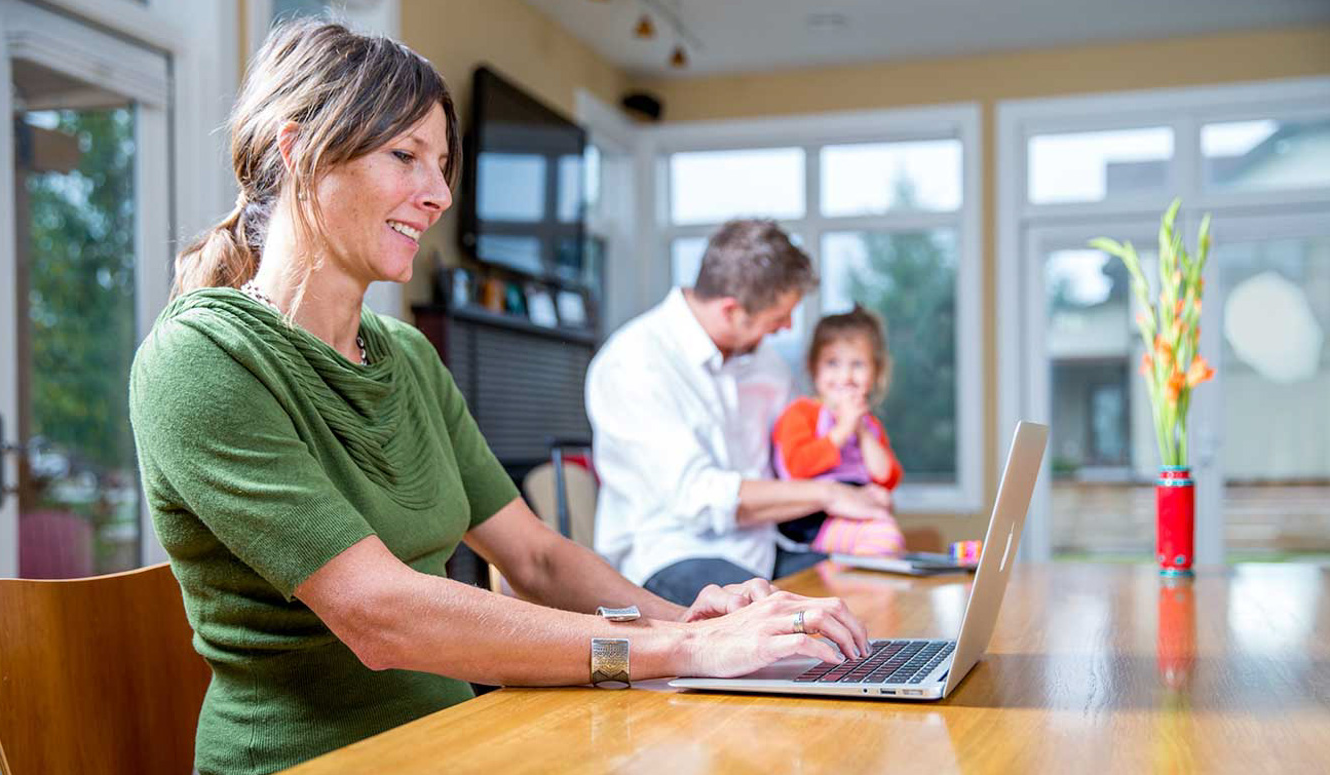 A woman working on a laptop at her kitchen table with her husband and daughter in the background.
