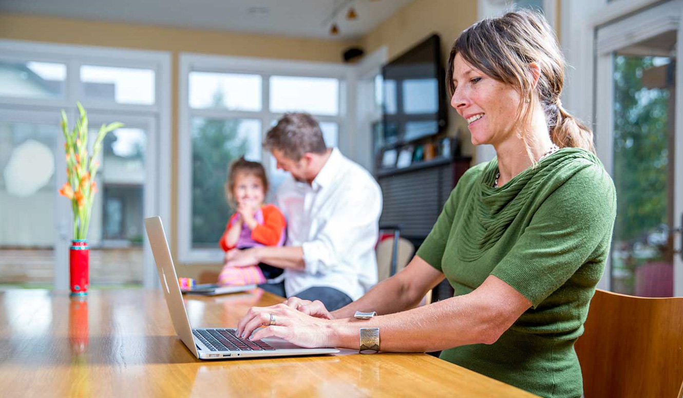 A woman working on a laptop at her kitchen table with her husband and daughter in the background.