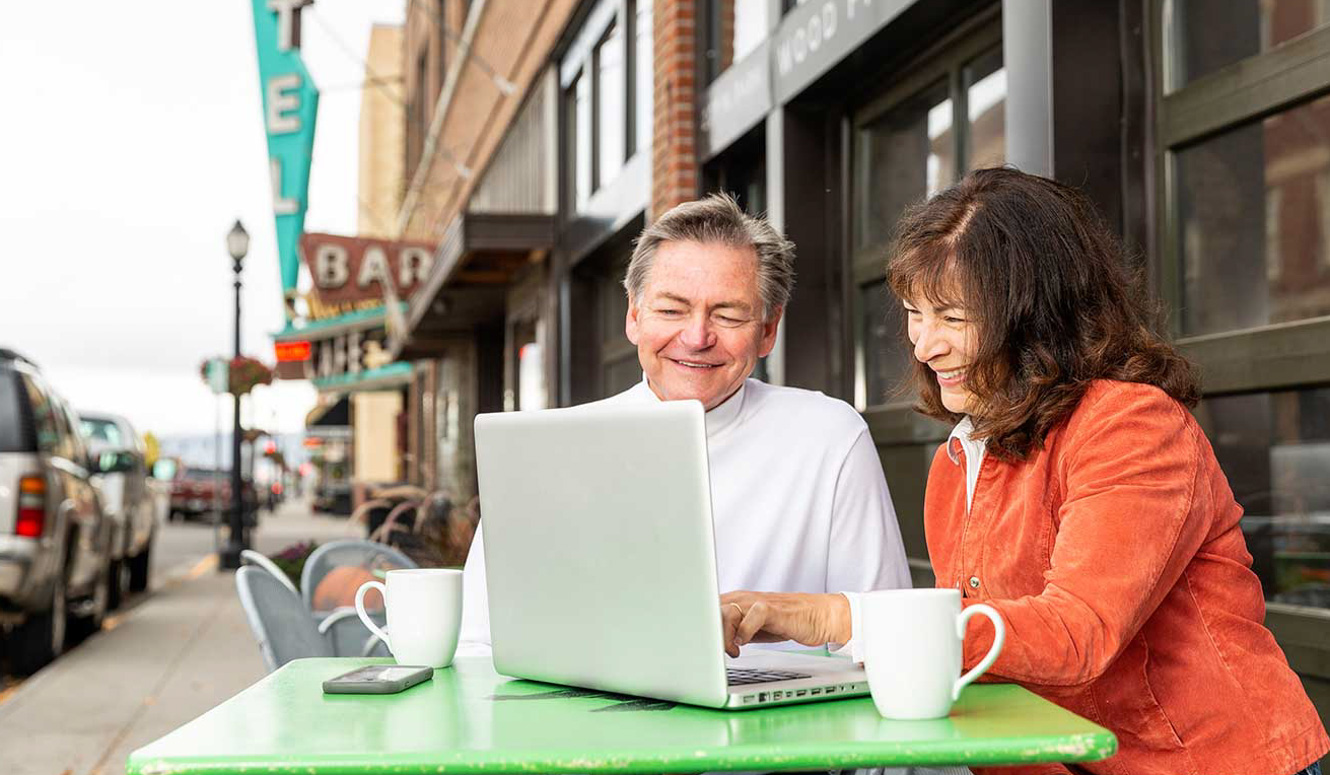A couple sitting at an outside cafe working on a laptop.