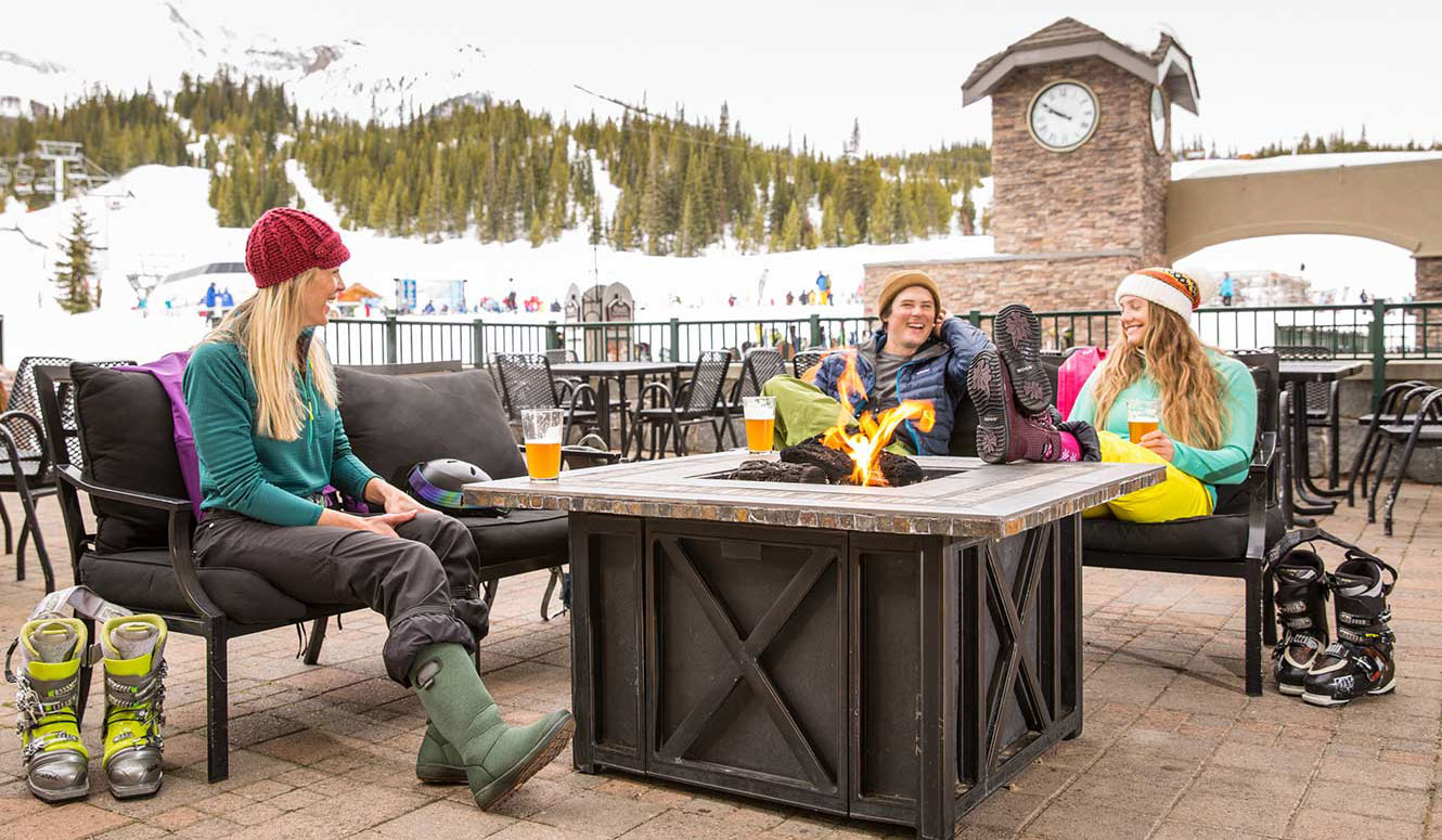 Three friends drinking beer around on an outdoor fireplace at Big Sky ski resort in Montana.
